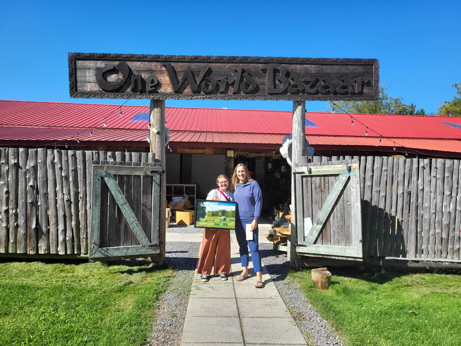 2 women standing outside the wooden gates of one world bazaar, one women is holding a painting of a hors